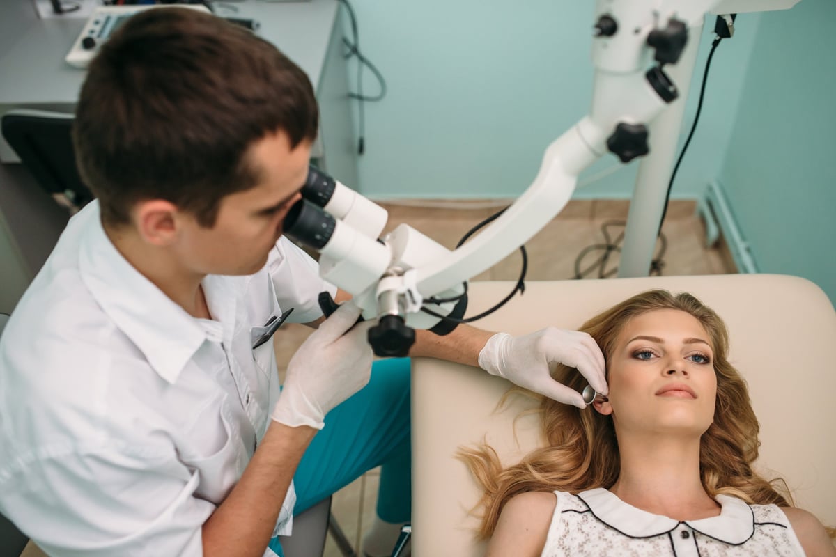 A healthcare professional uses a microscope to assess a patient's ear in a well-lit medical office.